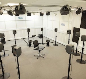 Listening room with multiple loudspeakers on metal stands and mounted on ceiling, with office chair in the middle.
