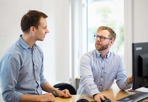 Two men in blue shirts talking to each other at desk